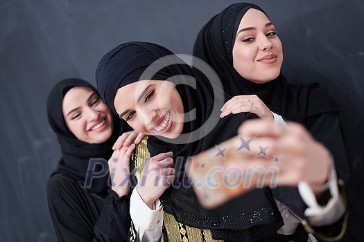 group of young beautiful muslim women in fashionable dress with hijab using mobile phone while taking selfie picture in front of black chalkboard representing modern islam fashion technology and ramadan kareem concept
