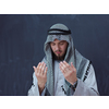 young arabian man in traditional clothes making traditional prayer to God, keeps hands in praying gesture in front of black chalkboard representing modern islam fashion and ramadan kareem concept