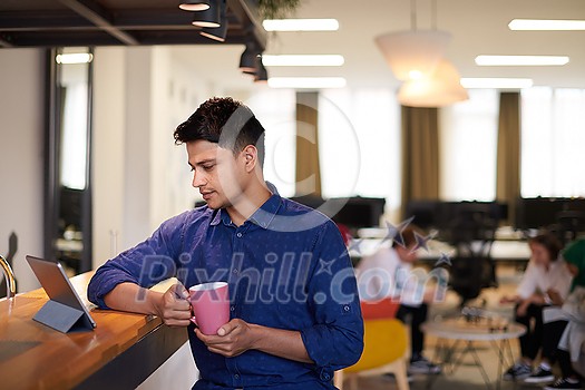 casual indian business man taking break from the work  using tablet computer while drinking tea in relaxation area of modern open plan startup office