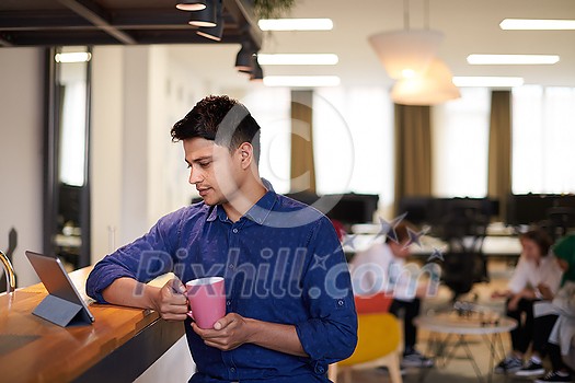 casual indian business man taking break from the work using tablet computer while drinking tea in relaxation area of modern open plan startup office casual indian business man taking break from the work using tablet computer while drinking tea in relaxation area of modern open plan startup office