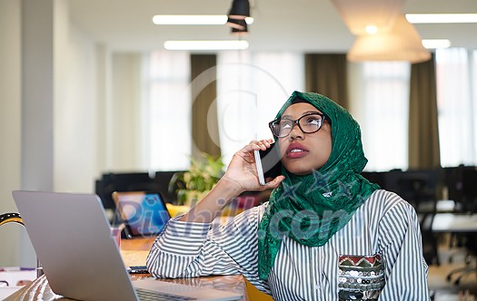 african muslim business woman wearing a green hijab using mobile phone while working on laptop computer in relaxation area at modern open plan startup office. Diversity, multiracial concept