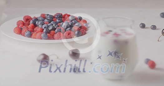 A glass with mix berries and milk. A plate filled raspberries, blueberries, cherries on a white table. The focus changes from the plate to the glass. Shallow depth of field. Slow motion video, 4K.