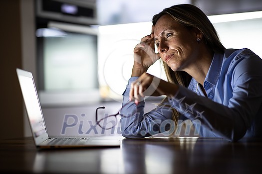 Pretty, middle-aged woman working late in the day on a laptop computer at home, running a business from home/working remotely for a corporation