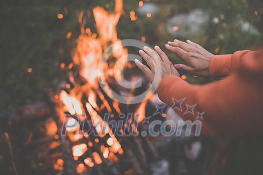 Young woman making fire while camping outdoors, in an alpine wilderness - warming up her hands near the fire
