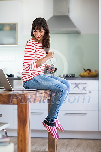 Successful young working from home. Portrait of happy entrepreneur working on a laptop computer while makin coffee. Doing business remotely from the comfort of her home.