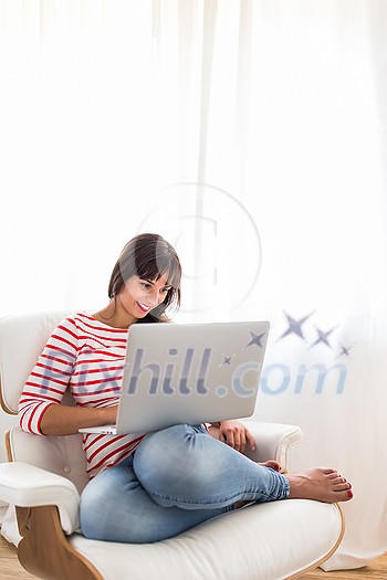 Successful young business woman sitting in a designer chair, working from home. Portrait of happy entrepreneur working on a laptop computer. Doing business remotely from the comfort of her home.
