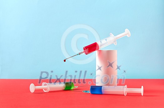 Creative healthcare composition of two plastic disposable syringes with blue and green drugs and one with drop of red liquid on a plaster cylinder on a duotone background, copy space.