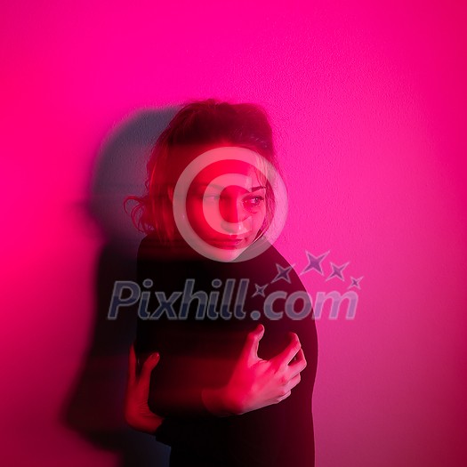 Depressed young woman in front of a wall lit by red light, lonely, anxious (color toned image)