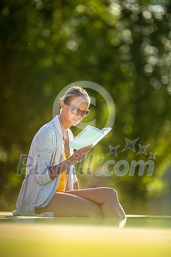 Young Beautiful Suntanned Woman wearing sunglasses relaxing next to a Swimming Pool  on a lovely Summer Day  - Reading a book