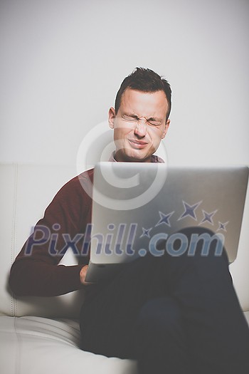 Exhausted young man with tired eyes, sitting on a sofa and trying to get some work done on his laptop computer Exhausted young man with tired eyes, sitting on a sofa and trying to get some work done on his laptop computer