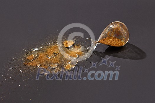 Old-fashioned aged crashed sandglass with glass shatters and golden sand on a black background with hard shadows, copy space.