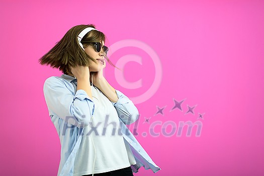 Gorgeous brunette lady in black glasses dancing and   listening music in headphones and singing on pink background.