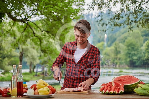 Man cooking tasty food on barbecue grill for outdoor french dinner party near the river on beautiful summer evening in nature