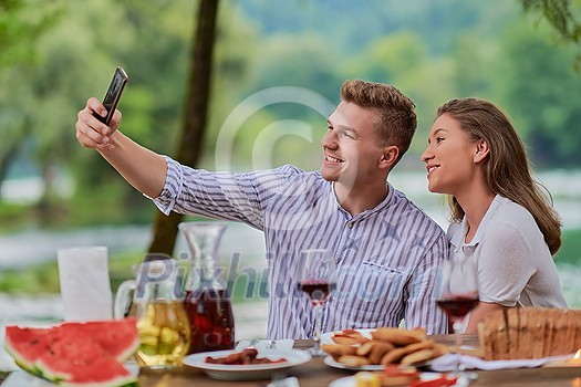 young happy couple taking selfie while having picnic french dinner party outdoor during summer holiday vacation near the river at beautiful nature