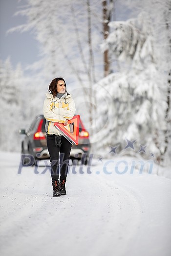 Pretty, young woman setting up a warning triangle and calling for assistance after her car broke down in the middle of nowhere on a freezing winter day