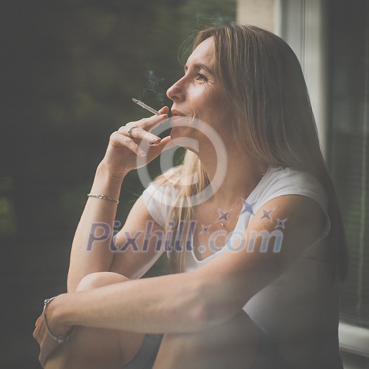 Mid-aged woman smoking a cigarette at home, getting her nicotine daily dose, unable to resist to her unhealthy habit Mid-aged woman smoking a cigarette at home, getting her nicotine daily dose, unable to resist to her unhealthy habit