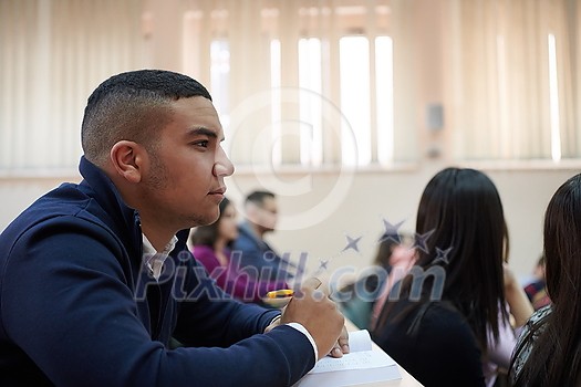 student taking notes while studying in high school. Satisfied young man looking at camera while sitting at desk in classroom. Portrait of college guy writing while completing assignment.
