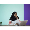 an afro girl wearing a hijab thoughtfully sits in her home office and uses a laptop
