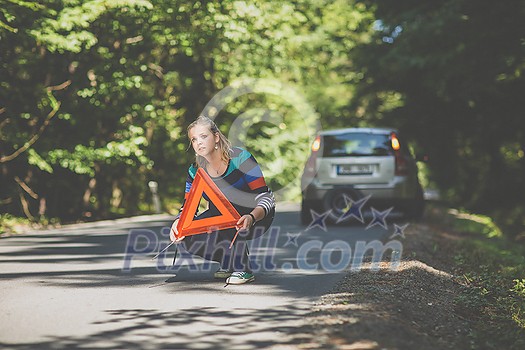 Pretty, young woman with her car broken down by the roadside, setting up the safety triangle, waiting for assistance