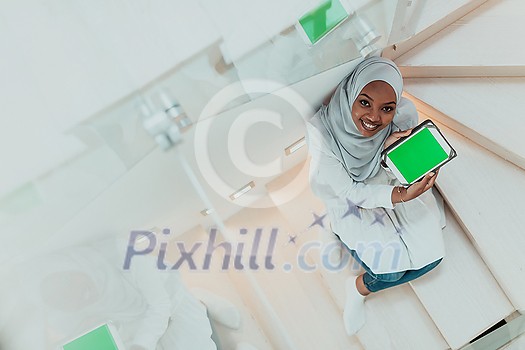 Young African modern Muslim woman using tablet computer while sitting on the stairs at home wearing hijab clothes top view. High-quality photo