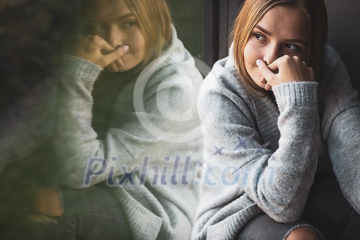 Depressed/anxious young woman sitting by a large window, feeling blue, sad, uncertain Depressed/anxious young woman sitting by a large window, feeling blue, sad, uncertain