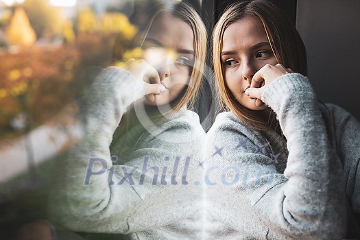 Depressed and anxious young woman sitting by a large window, feeling blue, sad, uncertain