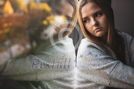 Depressed and anxious young woman sitting by a large window, feeling blue, sad, uncertain