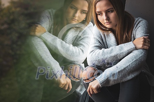 Depressed and anxious young woman sitting by a large window, feeling blue, sad, uncertain