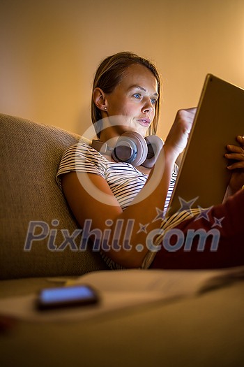 Pretty, young woman working at home in the evening - using her tablet computer as well as bluetooth headphones, writing down some ideas for tomorrow