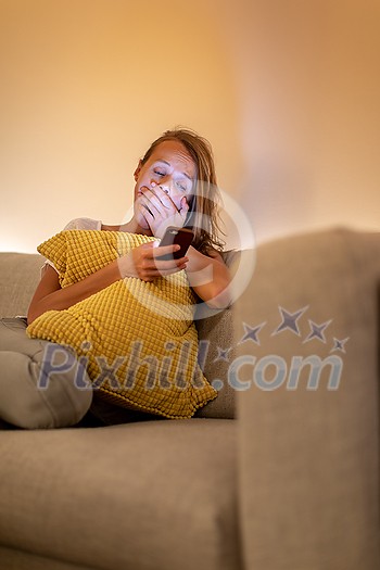 Cute young woman on a comfortable sofa in her modern apartment using her cell phone - staring at the blue lkight emitting device instaed of getting a good night sleep