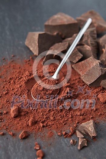 From above of organic dark bitter cocoa powder with teaspoon and heap of chocolate chunks on gray background From above of organic dark bitter cocoa powder with teaspoon and heap of chocolate chunks on gray background