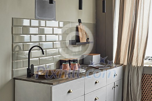 Small kitchen with white or grayish tiles wall with old fashion copper sink and modern faucet, tea and coffee containers, pink cups, and magazin on wooden counter. Country house kitchen concept.