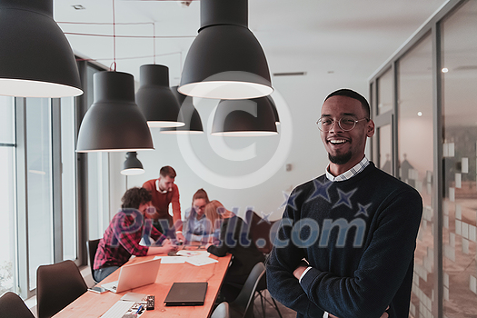 Portrait of happy Afro-American millennial male business owner in modern office. Businessman wearing glasses, smiling and looking at camera. Busy diverse team working in the background. Leadership concept. Head shot. High quality photo