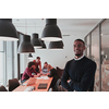 Portrait of happy Afro-American millennial male business owner in modern office. Businessman wearing glasses, smiling and looking at camera. Busy diverse team working in the background. Leadership concept. Head shot. High quality photo