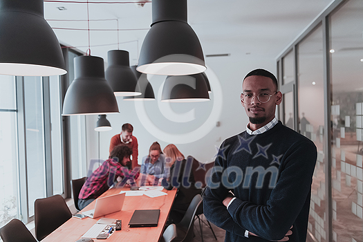 Portrait of happy Afro-American millennial male business owner in modern office. Businessman wearing glasses, smiling and looking at camera. Busy diverse team working in the background. Leadership concept. Head shot. High quality photo