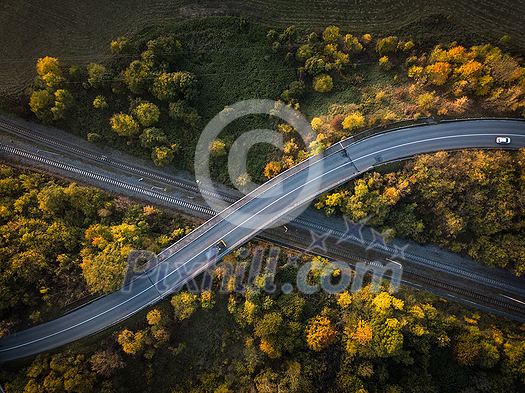 Winding road in autumn forest at sunset in mountains. Aerial view. Top view of beautiful asphalt roadway and orange trees. Highway through the woodland in fall. Trip in europe. Travel and nature