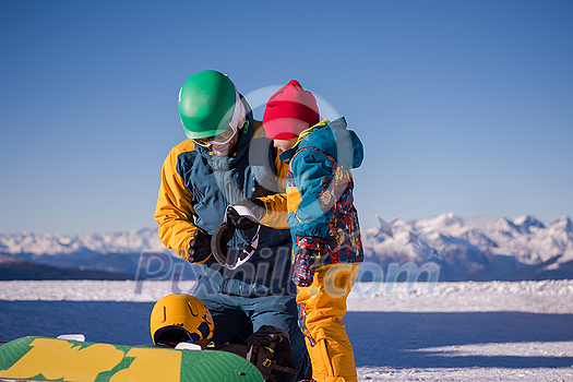 young happy father preparing his little son for the first time on a snowboard during sunny winter day at beautiful  ski resort