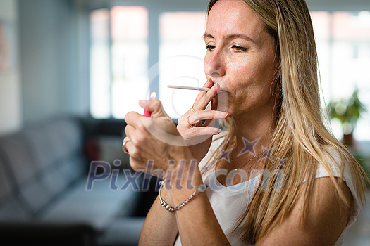 Mid-aged woman lighting a cigarette at home, getting her nicotine daily dose, unable to resist to her unhealthy habit Mid-aged woman lighting a cigarette at home, getting her nicotine daily dose, unable to resist to her unhealthy habit