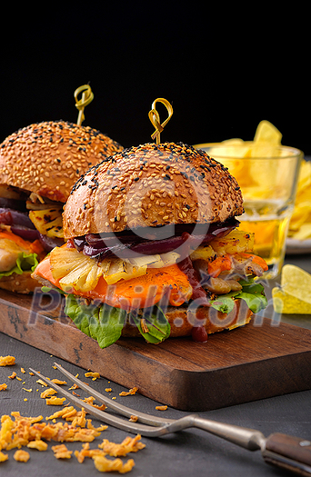 Tasty grilled chicken burger with lettuce, cheese, red onion and ananas served on cutting board on a black wooden table, with copyspace Tasty grilled chicken burger with lettuce, cheese, red onion and ananas served on cutting board on a black wooden table, with copyspace