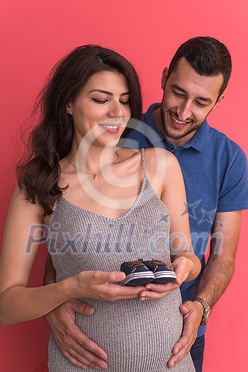 young  pregnant couple holding newborn baby shoes isolated on red background,family and parenthood concept