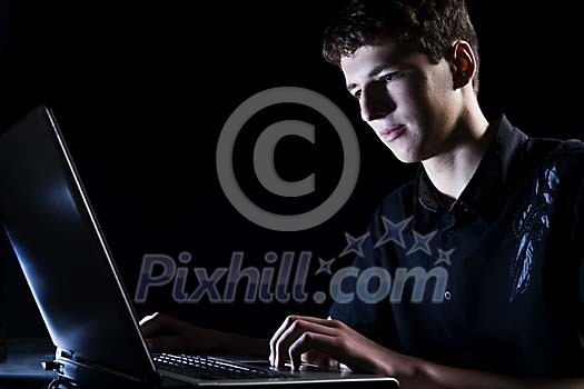 Teenage boy sitting with a laptop in a dark room Teenage boy sitting with a laptop in a dark room