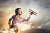 Image of little girl in pilots helmet playing with toy airplane against clouds background