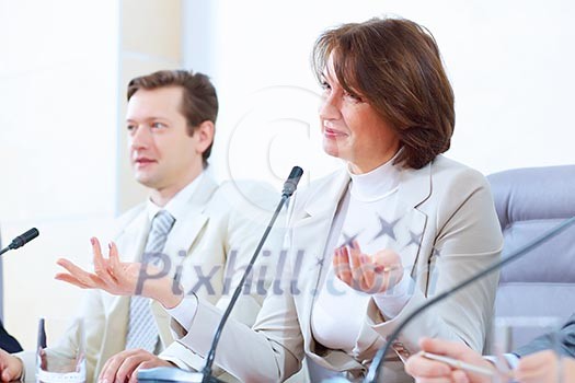Image of two businesspeople sitting at table at conference speaking in microphone Image of two businesspeople sitting at table at conference speaking in microphone