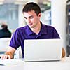 Handsome college student using his laptop computer in the campus common area/high school study room, absorbed (shallow DOF; color toned image)