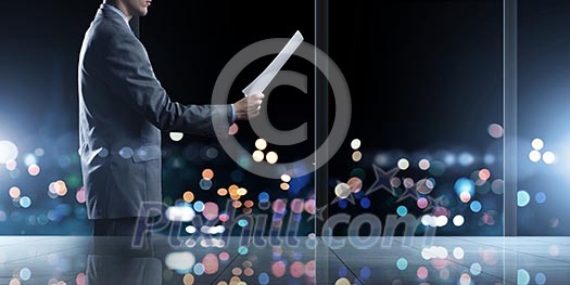 Businessman standing against office window reading documents Businessman standing against office window reading documents