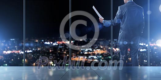 Businessman standing against office window reading documents Businessman standing against office window reading documents