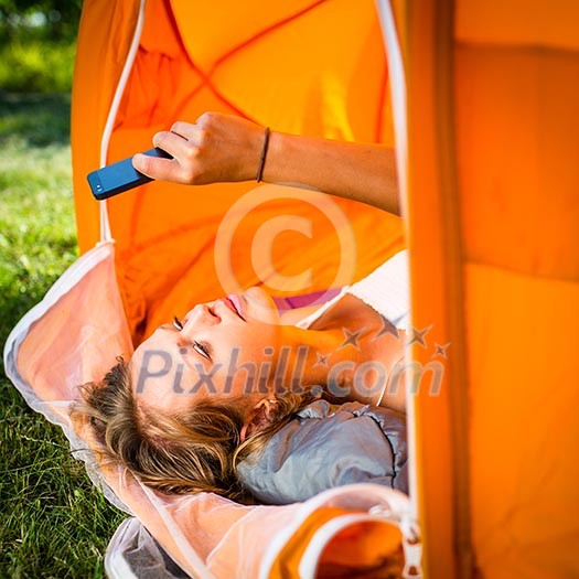 Pretty, young woman camping outdoors, lying in the tent in the morning, using her smart phone Pretty, young woman camping outdoors, lying in the tent in the morning, using her smart phone