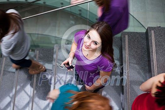 At the university/college - Students rushing up and down a busy stairway - confident pretty young female student looking upwards while listening to music on her mp3 player (color toned image) At the university/college - Students rushing up and down a busy stairway - confident pretty young female student looking upwards while listening to music on her mp3 player (color toned image)