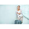 Pretty, young female passenger at the airport, waiting for her flight, wearing casual yet elegant clothes, arms folded, with a lovely smile and a big suitcase