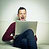 Sleepy young man sitting on a sofa and trying to get some work done on his laptop computer, yawning